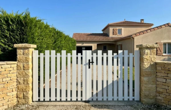 Portail battant blanc à lames verticales installé entre deux poteaux maçonnés avec des pierres